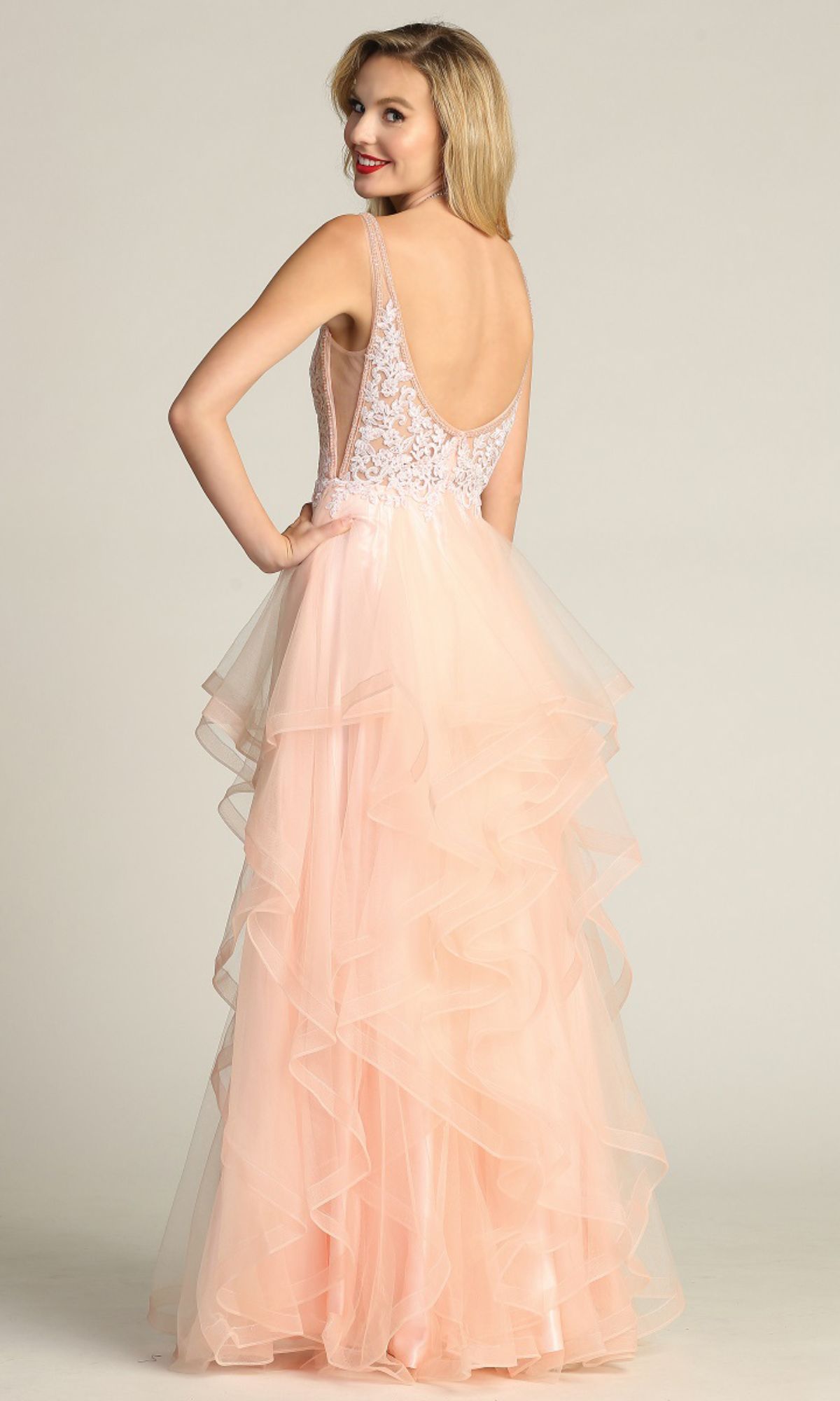 Woman in the Sheer-Sides Long Tiered Prom Dress (Chicas XR6006) with a sleeveless, backless blush pink lace bodice and layered tulle A-line skirt, standing and smiling over her shoulder.
